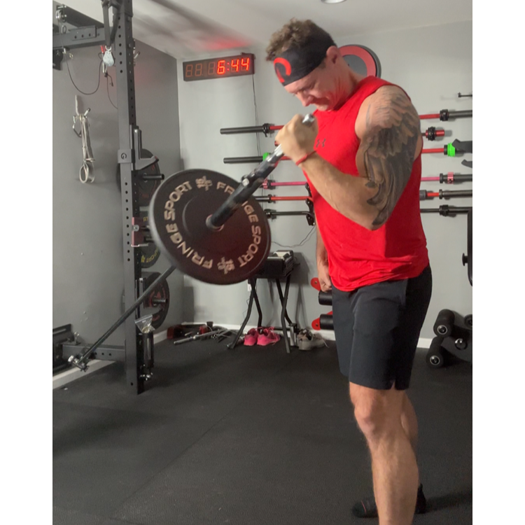 An athlete using a landmine bar for an exercise, wearing a red tank top and black shorts, with gym equipment in the background.