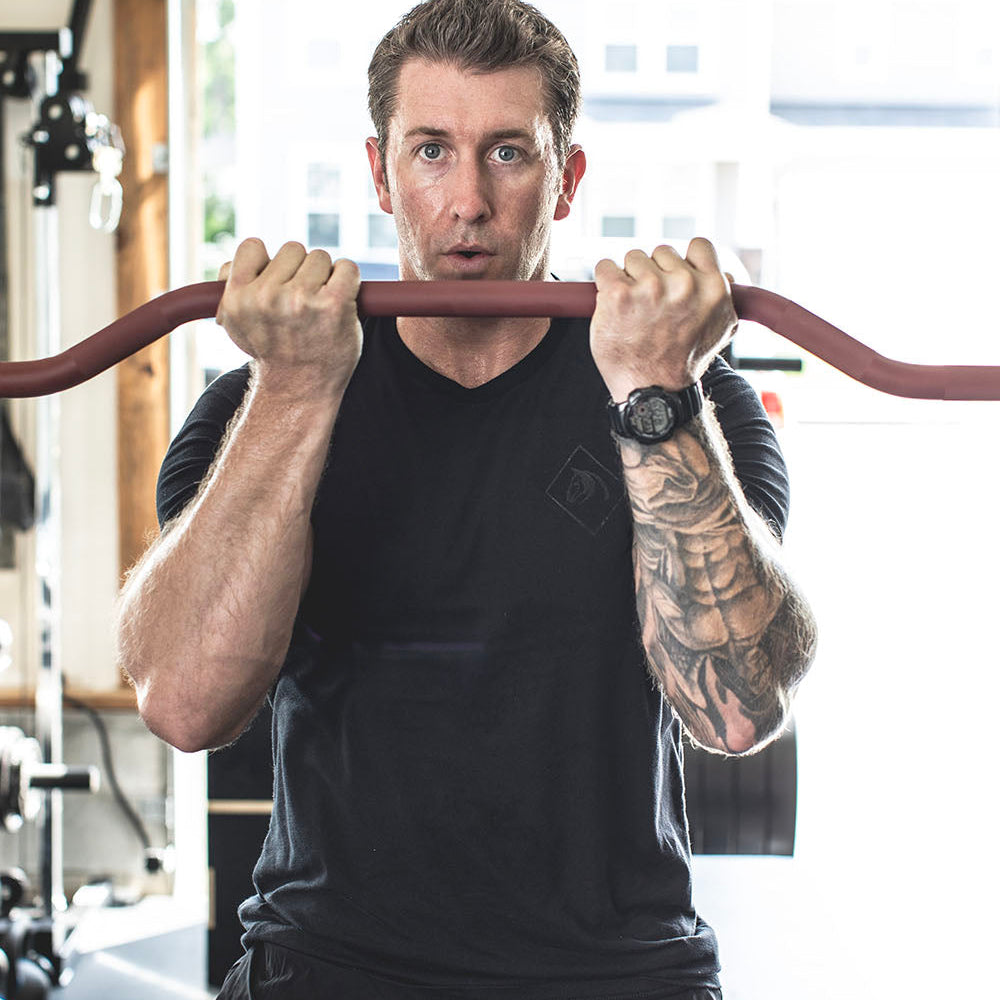 The Edge Bar is a mix between a shortened cambered bar and an EZ Bar. Perfect for enhanced bicep workouts, chest workouts, and more. This image presents the Edge Rackable Cambered Bar being used by a man for a bicep workout.